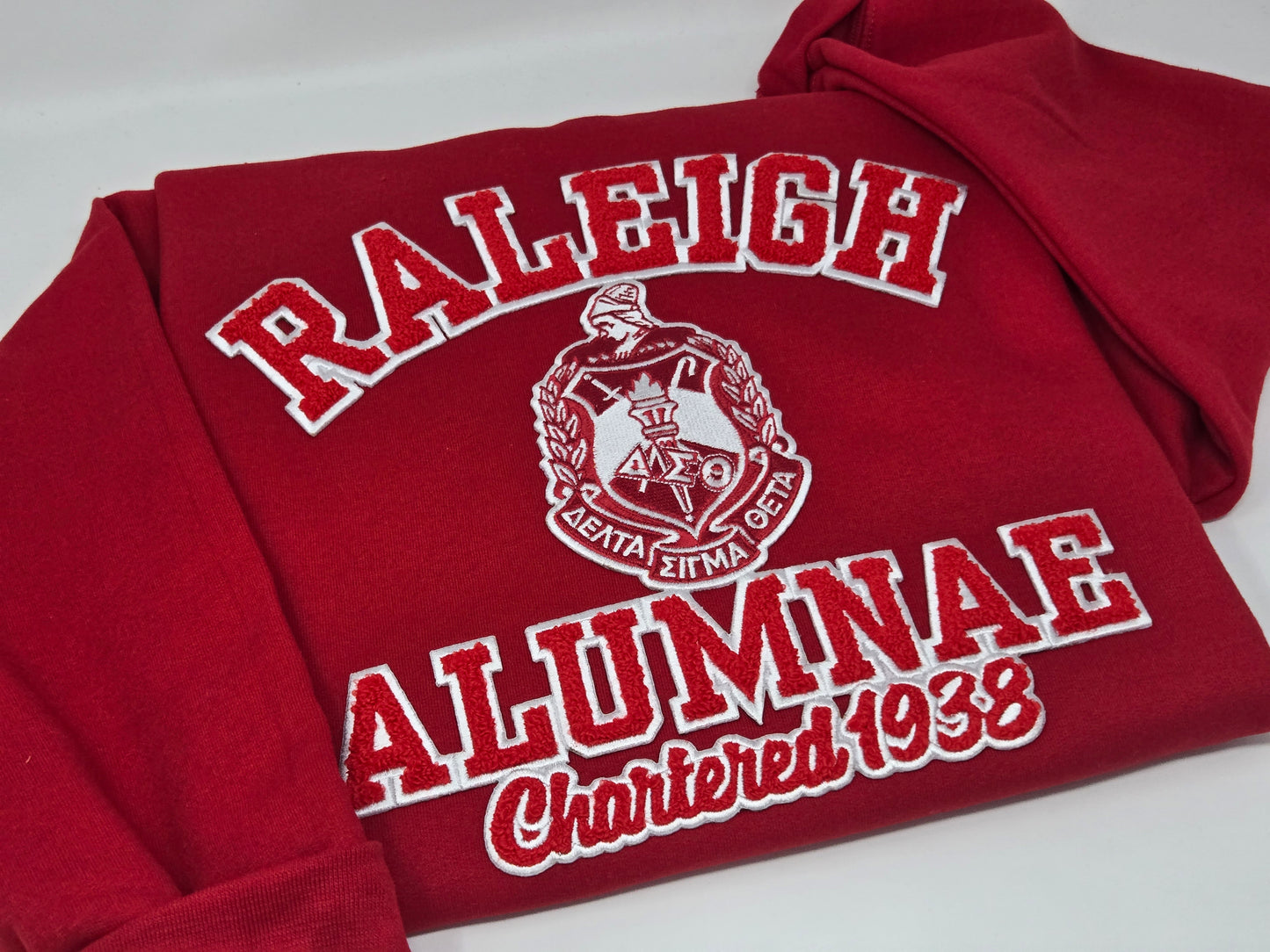 A red sweatshirt with 'RALEIGH ALUMNAE' and 'Chartered 1938' embroidered on it, representing the Delta Sigma Theta Sorority, Inc.