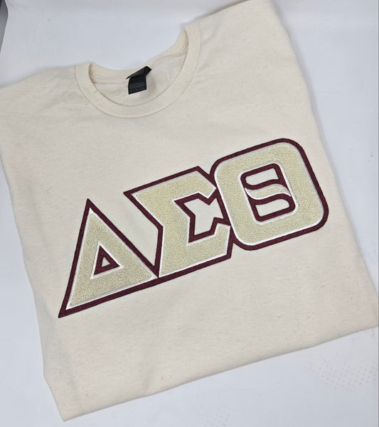 A folded cream-colored t-shirt with a chenille patch in crimson and cream letters.