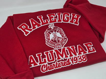 A red sweatshirt with 'RALEIGH ALUMNAE' and 'Chartered 1938' embroidered on it, representing the Delta Sigma Theta Sorority, Inc.