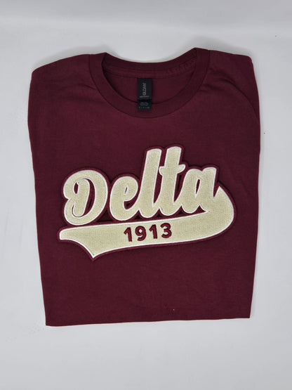 A maroon colored t-shirt with 'Delta 1913' embroidered in cream-colored chenille text on the front.